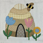 A handmade patchwork applique quilt design featuring a honeybee, a beehive, and three flowers on a textured off-white fabric background.