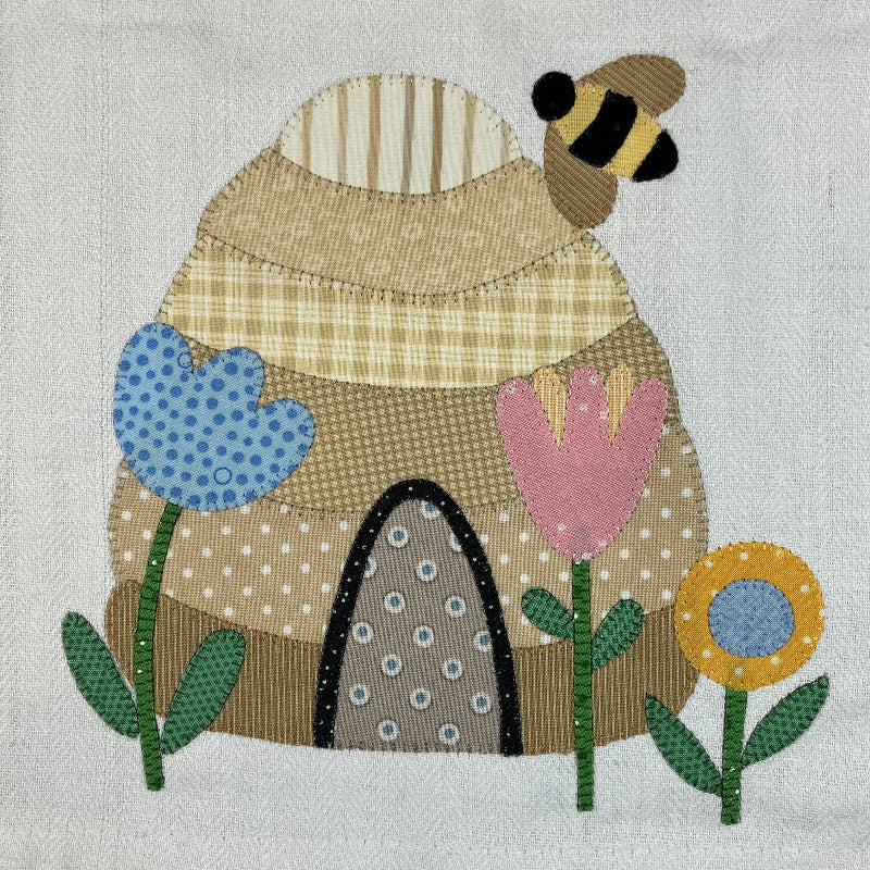 A handmade patchwork applique quilt design featuring a honeybee, a beehive, and three flowers on a textured off-white fabric background.