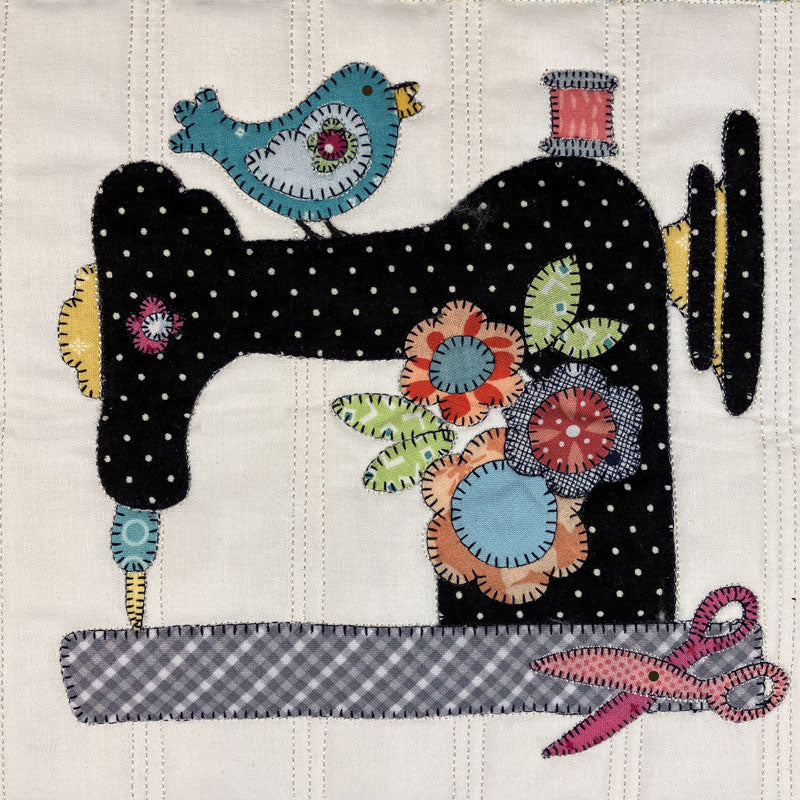 An applique design featuring a black sewing machine with white polka dots, a blue bird on top, colorful floral motifs, a gray-and-white gingham sewing needle path, and pink scissors on an off-white quilted background.
