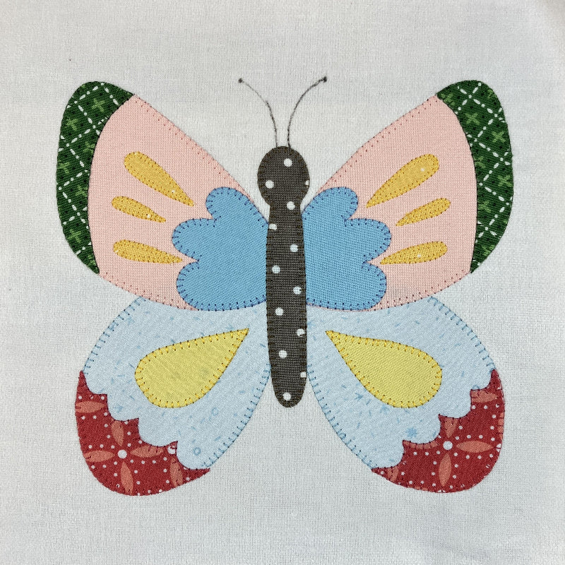 A colorful patchwork butterfly applique on a light gray fabric background. The butterfly features patterned wings in pink, blue, yellow, green, and red, with a dark gray body.