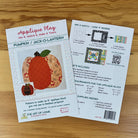 Front  and back covers for an applique block titled 'Applique Play' laid on a wooden surface. The front sheet shows a pumpkin design and the title 'Pumpkin / Jack-O'-Lantern,' while the back sheet explains how to mix and match patterns and lists included items and requirements.
