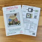 Front and back covers of an applique block pattern titled "Applique Play" by The Art of the Home, placed on a wooden surface. One sheet shows the front cover with a bee and flower applique design, and the other shows instructions and included colors.