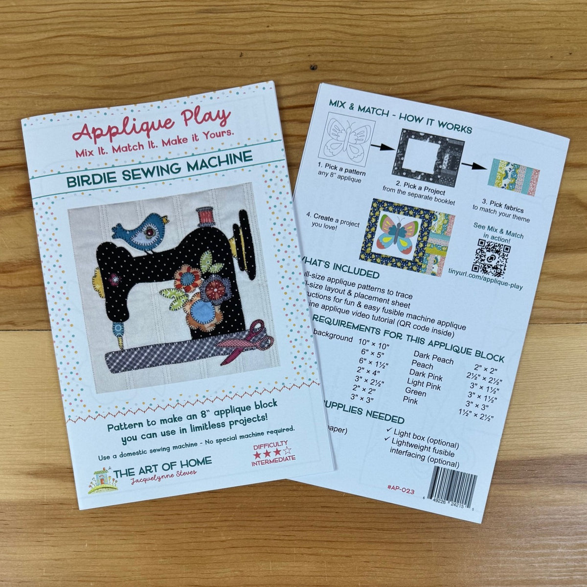 Front and back covers of 'Applique Play' for a 'Birdie Sewing Machine' by The Art of the Home, shown on a wooden tabletop. The front cover features a colorful applique design of a bird and flower, while the back cover displays step-by-step instructions and a requirements list.