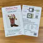 Two printed craft pattern cards for an applique block pattern titled 'Cowgirl Boot' by The Art of Home, placed on a wooden surface. One card shows the front design with a boot and floral applique illustration, and the other shows the back with instructions, included materials, and requirements.