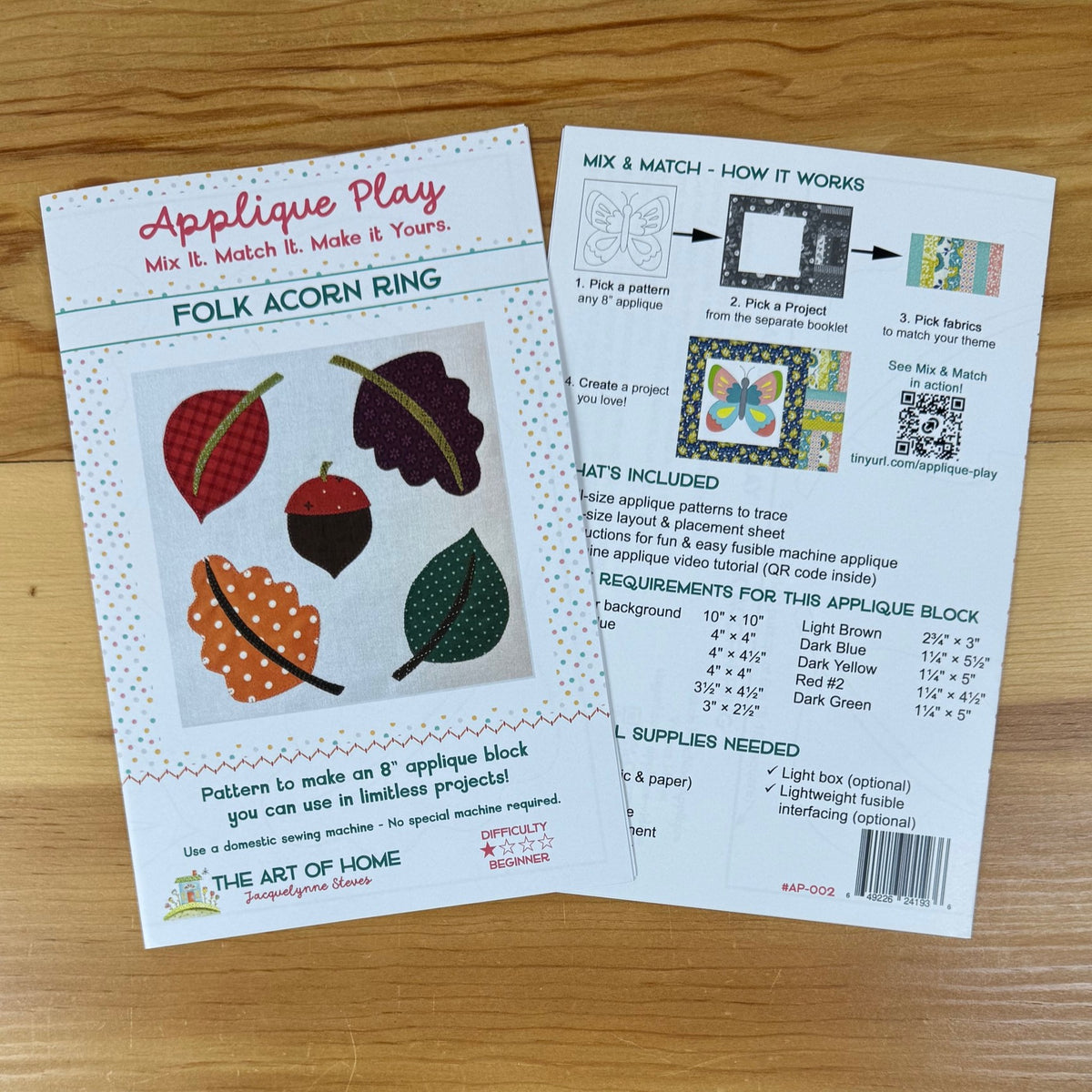 Front and back cover for a quilting applique pattern titled 'Applique Play' and 'Folk Acorn Ring' laid on a light wood surface. The front card shows illustrated acorns and the product name, while the back card shows instructions, included materials, and a requirements table.