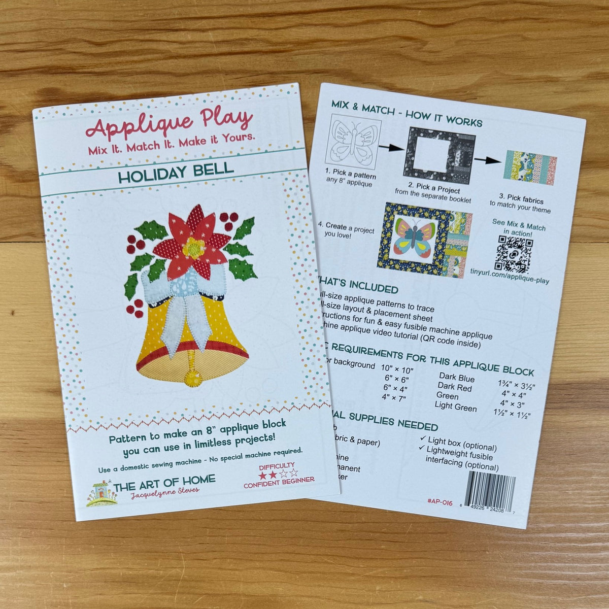 Front and back covers for an applique pattern titled 'Holiday Bell' by The Art of the Home, placed on a wooden surface. One card shows the front with a colorful applique design of a bell and poinsettia, and the other shows the back with instructions, included materials, and a requirements table.