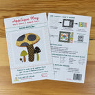Front and back covers for 'Applique Play' mushroom-themed patterns, shown on a light wood surface. The front card shows colorful mushroom applique designs and product title text, while the back card includes instructions, measurements, included fabrics, and a barcode.