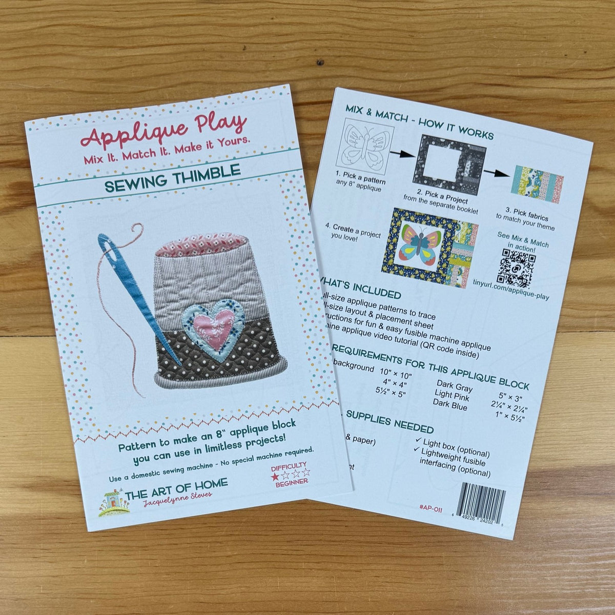 Front and back covers for an applique pattern titled 'Applique Play' by The Art of Home, placed on a light wood surface. One sheet shows the front cover with a sewing thimble illustration and product title, and the other shows instructions and included materials.
