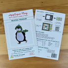Front and back covers of pattern for making a Skating Penguin applique block, placed on a light wood surface. One sheet shows the front cover with a penguin illustration and title text, and the other shows the back with instructions, included materials, and a QR code.