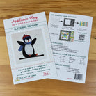 Front and back covers for an applique block featuring a penguin design, placed on a wooden surface. One sheet shows the front cover with the title 'Applique Play' and 'Sledding Penguin,' while the other shows instructions and included colors/materials.