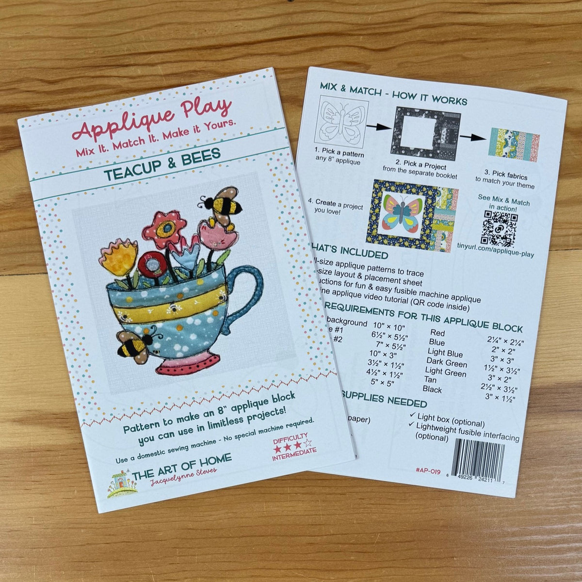Front and back covers for the Teacup & Bees Applique Play pattern set, one showing the front cover with colorful teacup illustrations and the other showing the back with instructions, included items, and requirements, photographed on a wooden surface.
