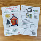 Front and back covers for The Art of Home 'Applique Play' Umbrella Beach Shack pattern, showing the front cover with an illustrated beach shack and the back with instructions, materials, and requirements, photographed on a wooden surface.