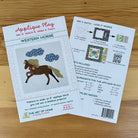 Two printed craft pattern sheets for an applique block titled 'Western Horse' by The Art of Home, placed on a light wood surface. The front sheet shows a horse applique design and product details, while the back sheet explains how to mix and match colors and includes included fabrics and requirements.