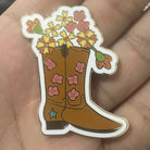 A cowboy boot enamel needle minder with pink flowers and yellow flowers growing out of the boot, held in a person's hand.
