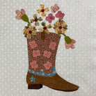 A quilted fabric applique of a brown cowboy boot filled with a bouquet of colorful floral patches, set against a light beige patterned fabric background.