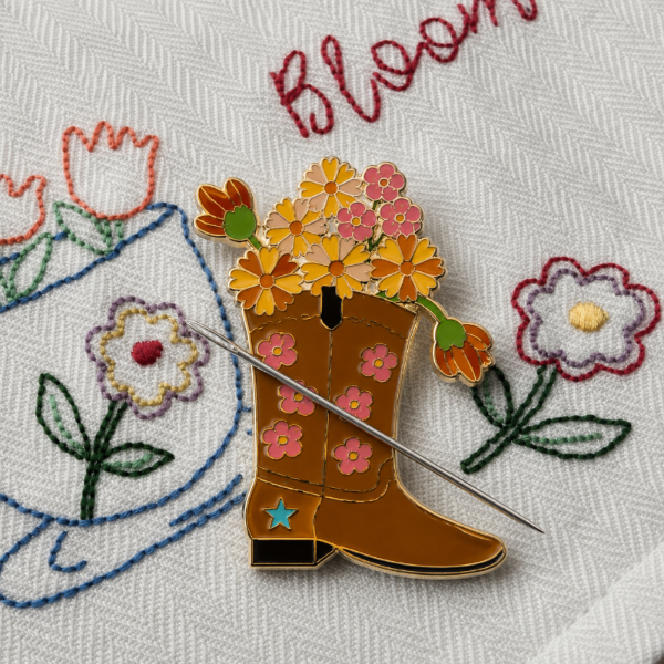 A decorative enamel needle minder shaped like a brown cowboy boot filled with colorful flowers, placed on white embroidered fabric with floral stitching and the word 'Bloom' visible.