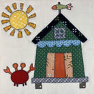 A square fabric applique design on an off-white background, featuring a green house with a dark roof, blue scalloped trim, patterned door panels, a yellow sun with orange rays, and a red crab.