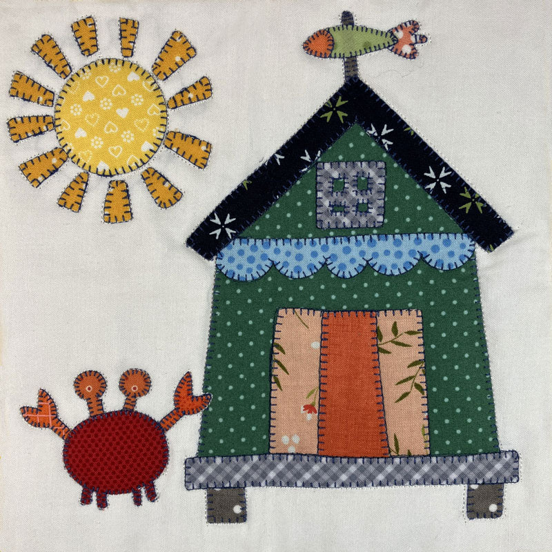 A square fabric applique design on an off-white background, featuring a green house with a dark roof, blue scalloped trim, patterned door panels, a yellow sun with orange rays, and a red crab.