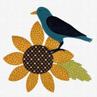 A stylized patchwork applique design of a blue bird perched on a yellow sunflower with a patterned brown center and green leaf, shown on a white textured fabric background.