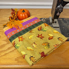 A folded piece of fabric with an autumn leaf pattern and a purple floral border, displayed on a wooden table next to a vintage sewing machine with a small pumpkin decoration.