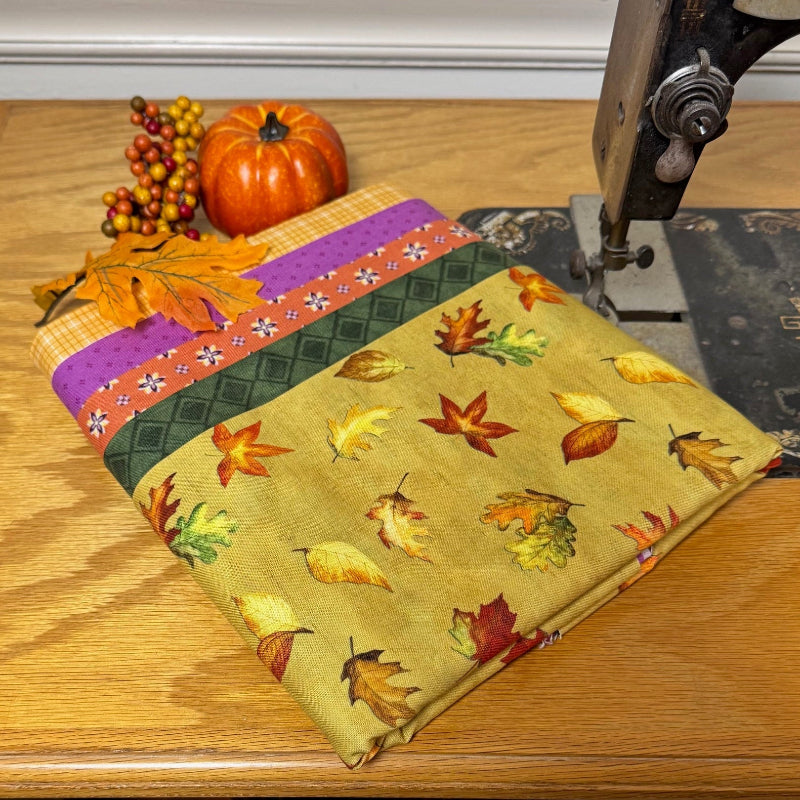 A folded piece of fabric with an autumn leaf pattern and a purple floral border, displayed on a wooden table next to a vintage sewing machine with a small pumpkin decoration.