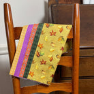 A close-up of a woven chair seat with a yellow and orange autumn leaf patterned fabric draped over the backrest, set in a home interior next to a wooden cabinet.