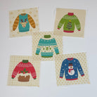 Five small square fabric patches featuring different colorful Christmas sweater designs, arranged on a white tabletop. 