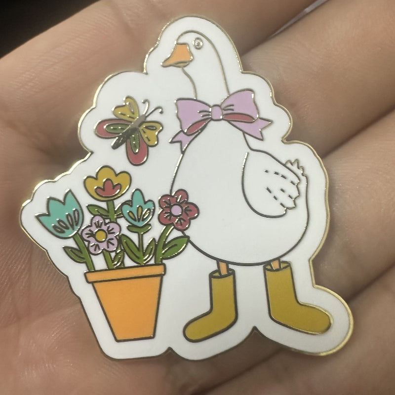A hand holding an enamel needle minder featuring a white goose wearing a pink bow and yellow rain boots, next to a potted flower with a butterfly.