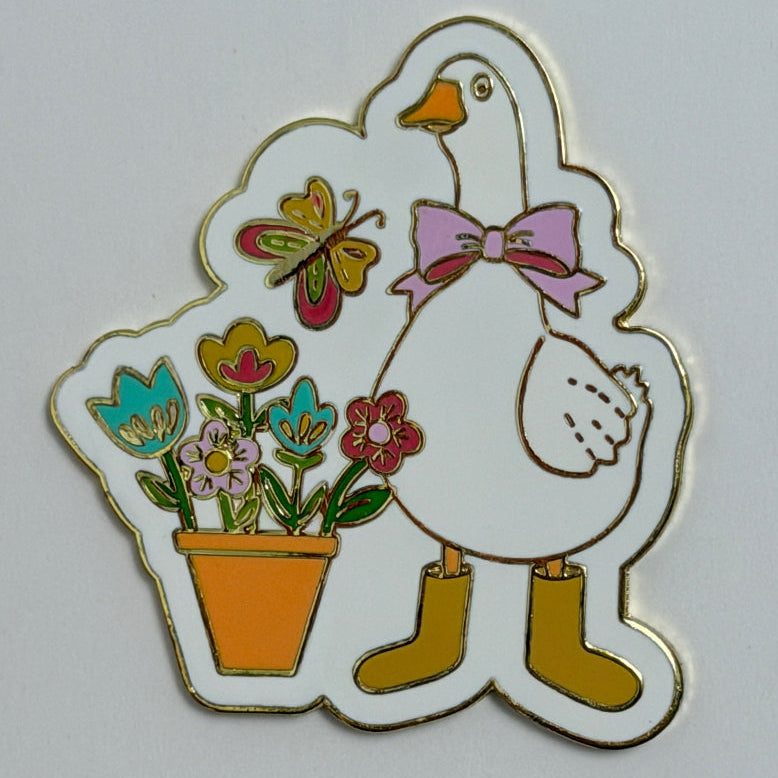 A enamel needle minder featuring a white goose wearing a pink bow and yellow boots, standing next to a potted flower arrangement with a butterfly, on a plain light background.