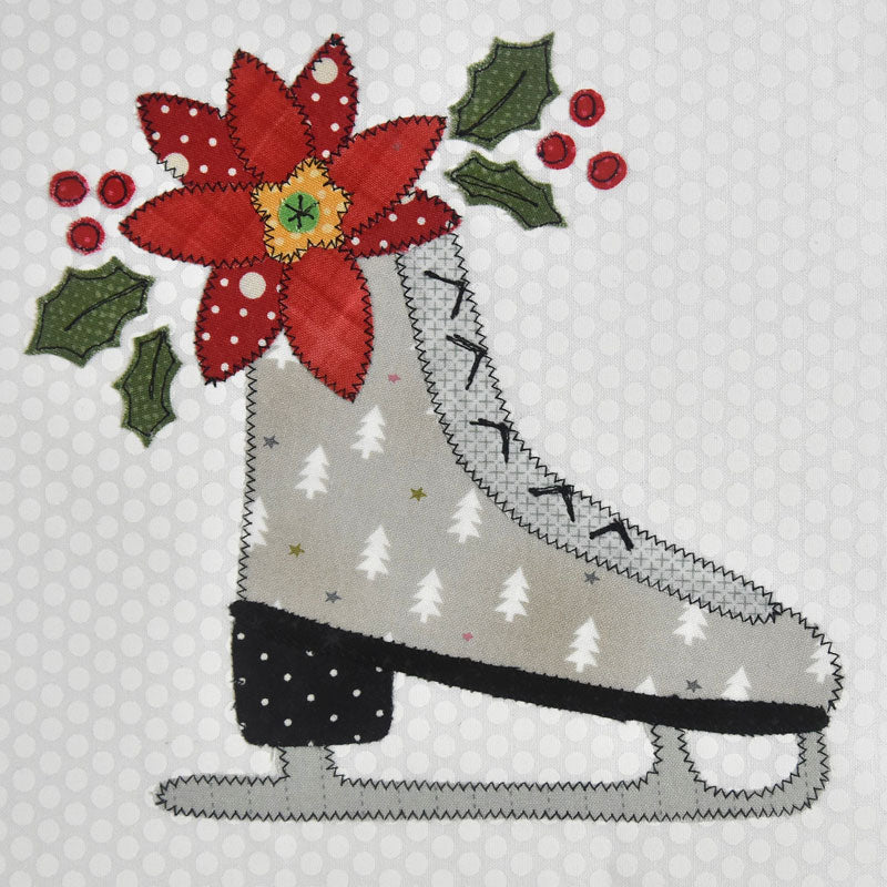 A festive applique or design of a light gray ice skate decorated with white pine trees, a red poinsettia flower, green holly leaves, and red berries on a white patterned fabric background.