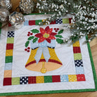 A square quilted fabric placemat or table topper featuring a festive Christmas design with two yellow bells, a red poinsettia, green holly leaves, and a multicolor patchwork border, displayed on a wooden tabletop with holiday greenery and silver ornaments.