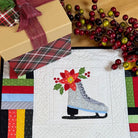 A square quilted fabric placemat or runner featuring a stitched gray ice skate with a red poinsettia and green leaves on a white center panel, surrounded by multicolored patchwork borders. It is displayed on a wooden table with holiday decorations, including a wrapped gift box and red berry branches.