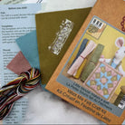 A Little Mice Wool Mix Felt Craft Kit package lying on top of instruction sheets and thread spools. The package is tan with a photo of a white rabbit holding a quilt, and the kit includes multicolored thread, pattern templates, and fabric pieces.