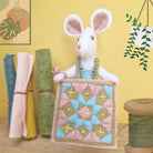 Stuffed mouse holding a quilt with fabric rolls and spools in the background
