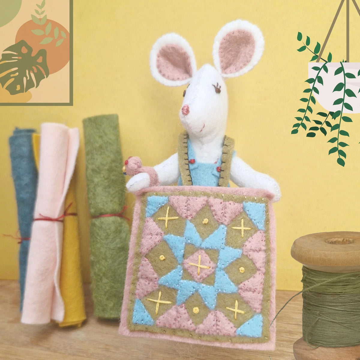 Stuffed mouse holding a quilt with fabric rolls and spools in the background