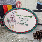 An oval embroidered Christmas ornament featuring a goose wearing a red and white striped scarf next to the text 'Have yourself A Merry Little Christmas' in green and blue thread. The ornament has a green polka-dotted border with red scalloped edges and hangs from a red-and-white candy cane loop.