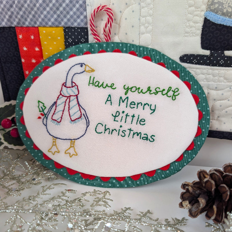 An oval embroidered Christmas ornament featuring a goose wearing a red and white striped scarf next to the text 'Have yourself A Merry Little Christmas' in green and blue thread. The ornament has a green polka-dotted border with red scalloped edges and hangs from a red-and-white candy cane loop.