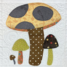 A quilted fabric applique design of three stylized mushrooms on a white quilted background. The main mushroom has a yellow-orange patterned cap with dark gray spots and a brown polka-dotted stem.