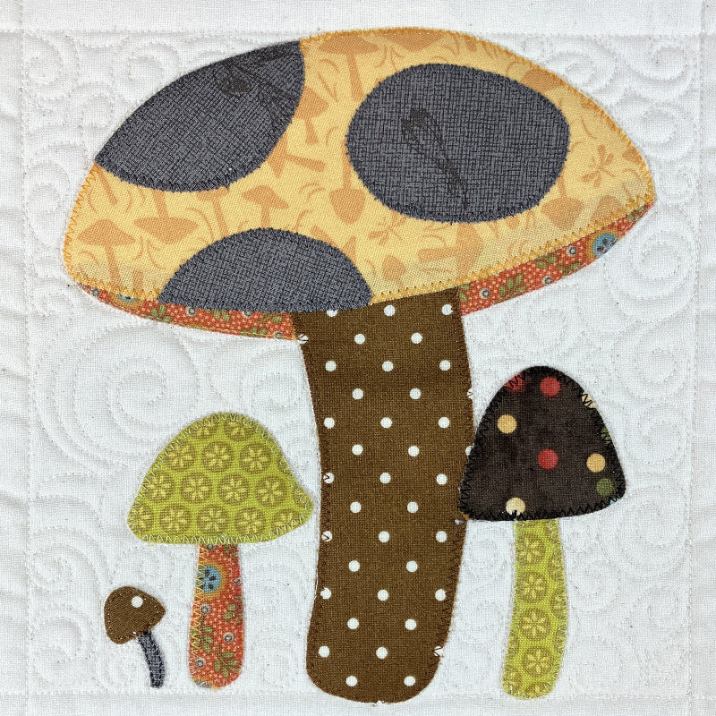 A quilted fabric applique design of three stylized mushrooms on a white quilted background. The main mushroom has a yellow-orange patterned cap with dark gray spots and a brown polka-dotted stem.