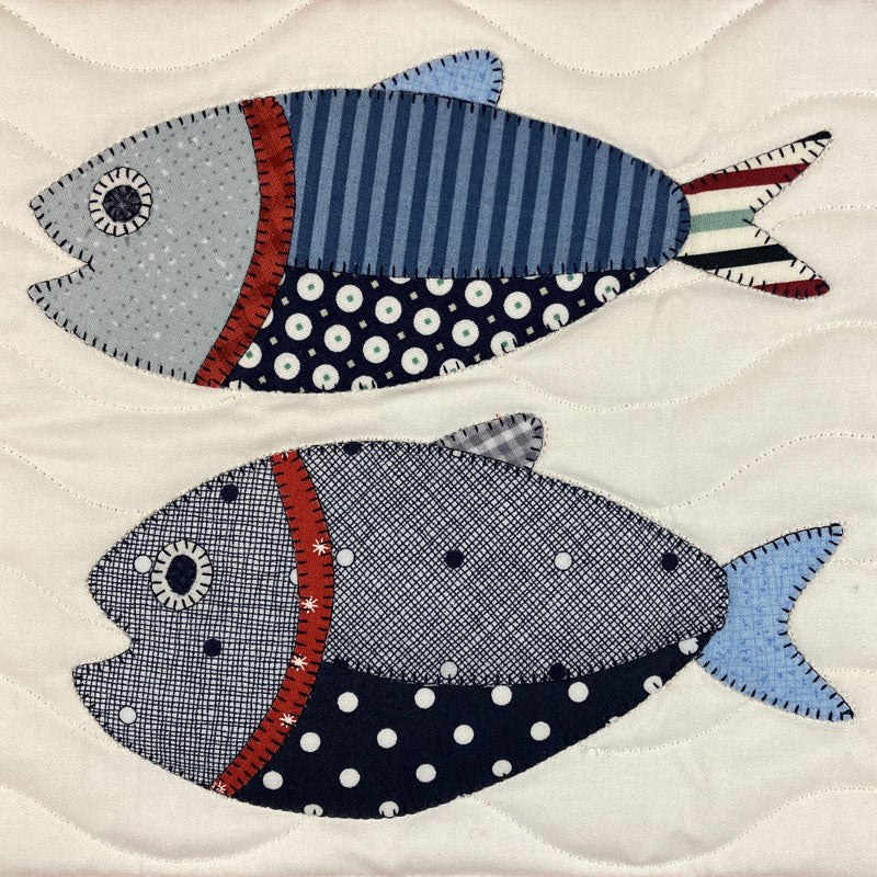 Two stylized fish appliqués on a quilted cream fabric background. The top fish is light blue and navy with red and white striped accents, and the bottom fish is gray and navy with white polka dots and a red stripe.