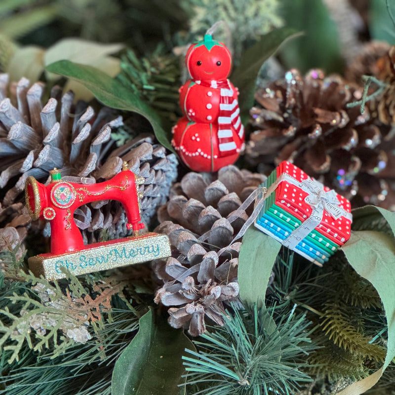 Decorative Christmas ornaments including a red sewing machine, tomato pincushion snowman, and fat quarter bundle on a bed of pine cones and greenery.