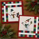Two square Christmas-themed placemats with quilted borders and pre-printed panel snowmen, arranged on a wooden surface with pine branches and red berries as styling props.