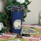 A navy blue insulated tumbler with a clear lid and a colorful bird graphic sits on a patchwork quilted table runner, surrounded by white flowers and green leaves.