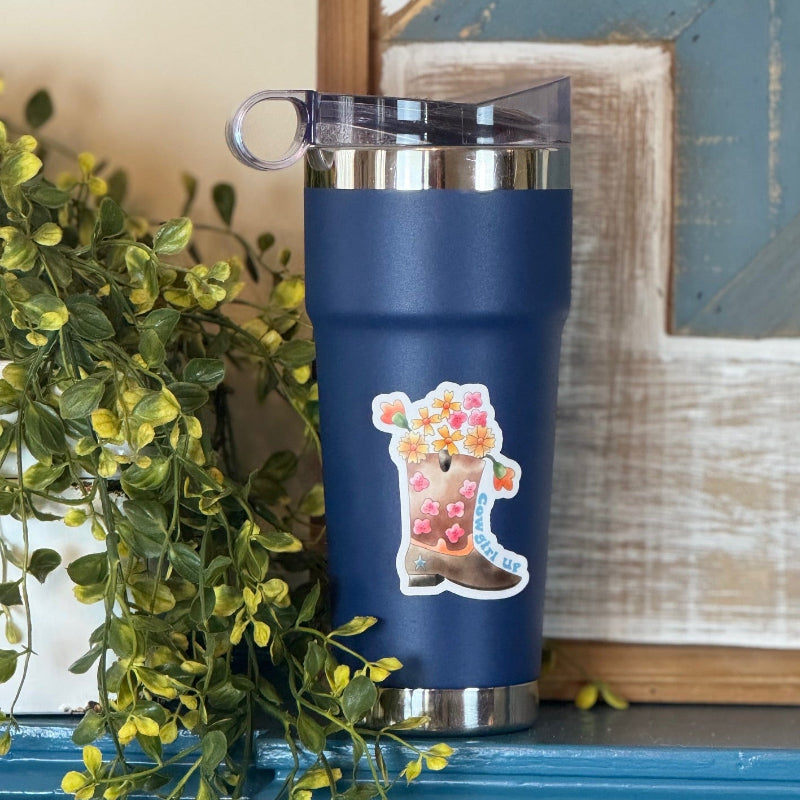 A navy blue insulated tumbler with a clear lid sits on a blue surface next to green trailing plants. The tumbler has a decorative sticker of a boot filled with flowers and the word 'PRAIRIE'.