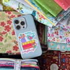 A large dark brown box filled with multiple stacks of colorful fabric bundles and quilts, photographed from above on a table. A light blue smartphone with a decorative case rests on top of the fabrics.