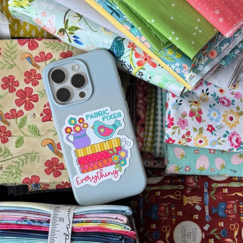 A large dark brown box filled with multiple stacks of colorful fabric bundles and quilts, photographed from above on a table. A light blue smartphone with a decorative case rests on top of the fabrics.