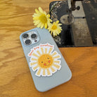 A light blue phone case with a smiling yellow sun sticker and the words 'Good Day Sunshine!' in red script, resting on a wooden table next to yellow daisies and a vintage sewing machine.