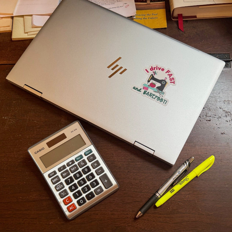 Laptop computer on desk with a sewing machine sticker that says, I Drive Fast and Barefoot.