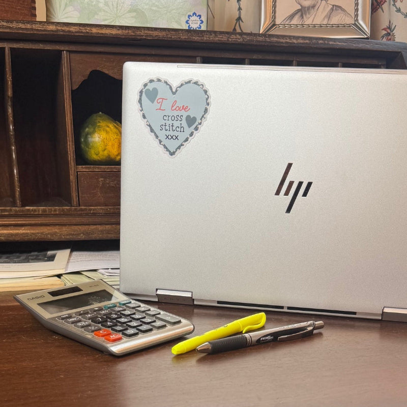 Silver laptop on antique desk with I Love Cross Stitch Sticker and calculator and pens in foreground.