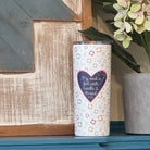 A white insulated tumbler with a colorful quilt star pattern and a purple heart graphic that reads 'My heart is Fed with Needle & Thread', displayed on a blue wooden mantel next to a potted plant.